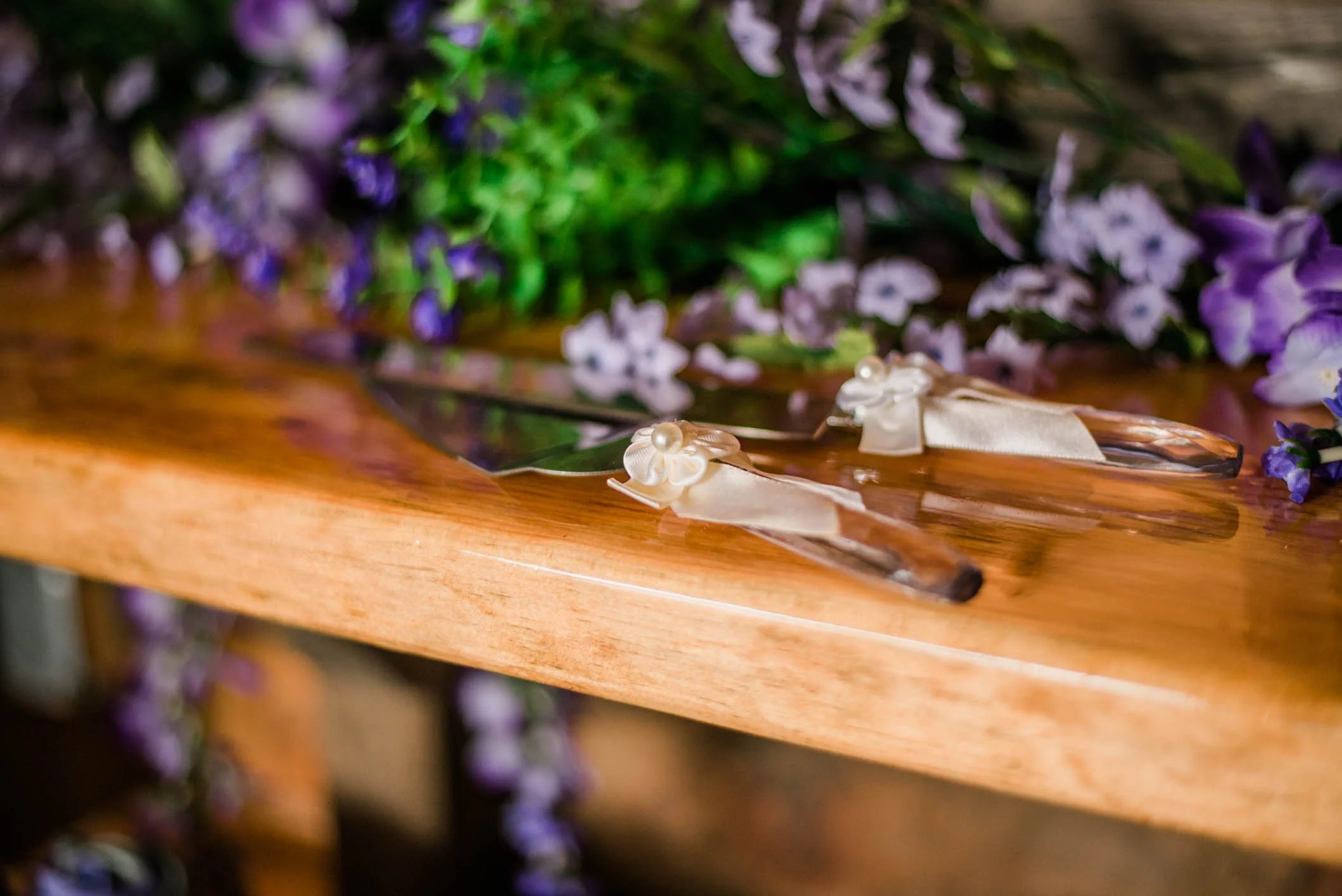 A pair of decorative knives with cream-colored ribbons and pearl embellishments, placed on a polished wooden table surrounded by purple and white flowers.