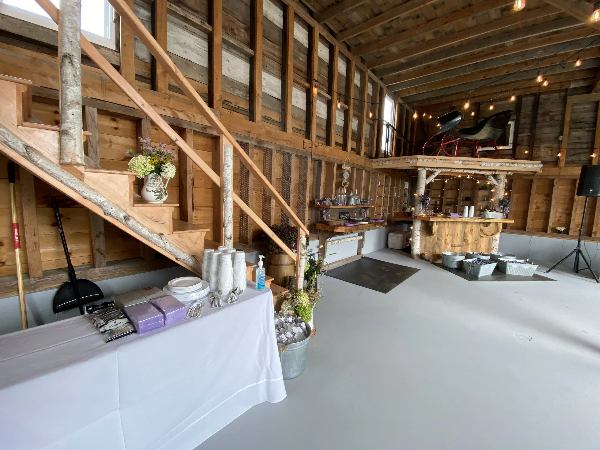 Interior of a rustic barn decorated for an event with string lights, a table holding cups, plates, and utensils, a bar area, and vintage sleds on a loft