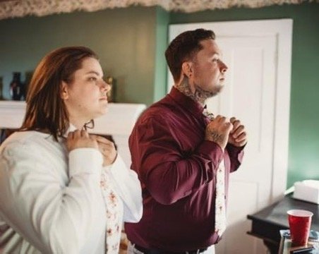 A man and woman standing indoors, holding necklaces near their necks, looking serious or contemplative.