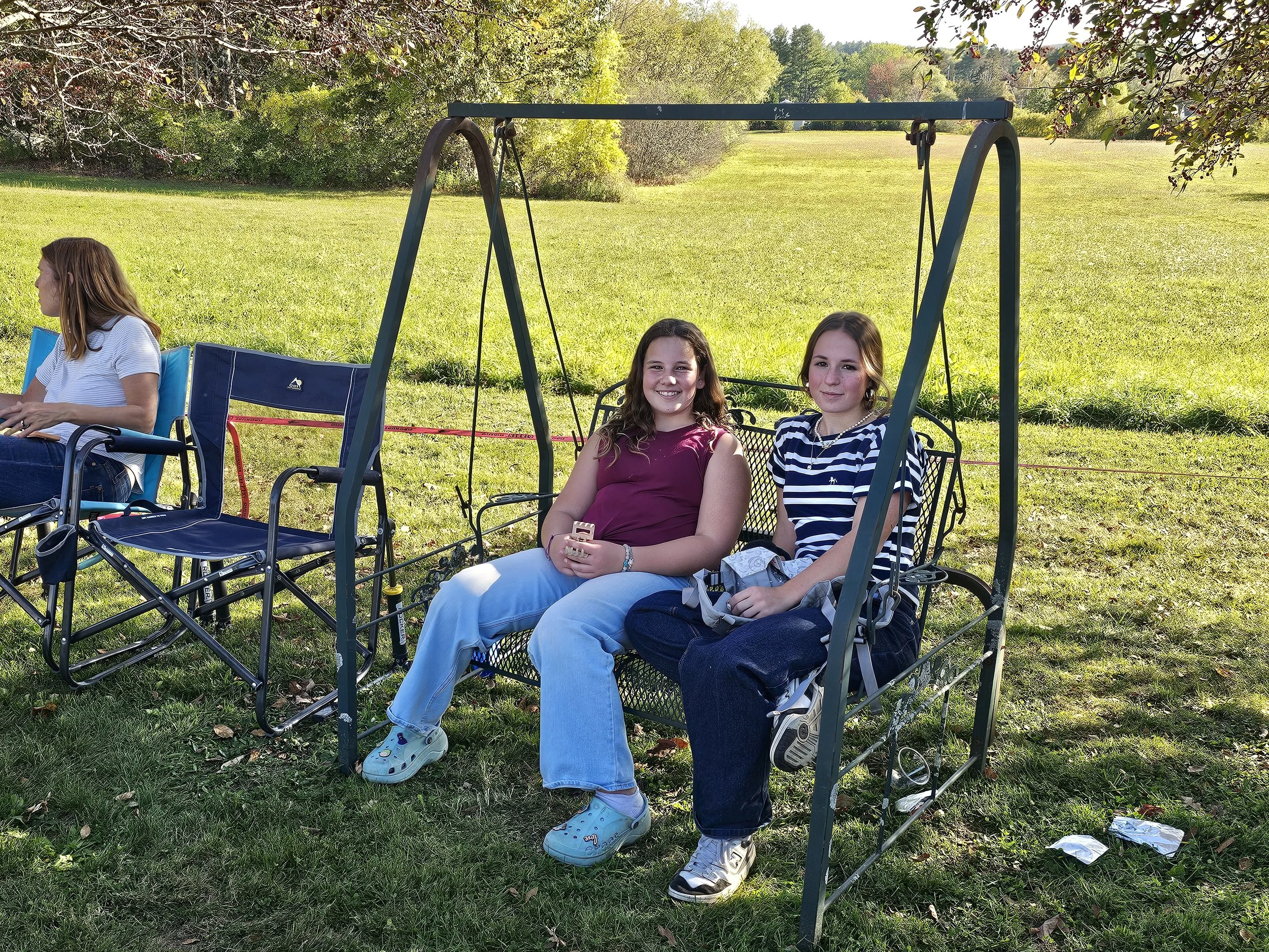 Four girls sitting on a swing set in a grassy park, under trees with green foliage, on a sunny day.