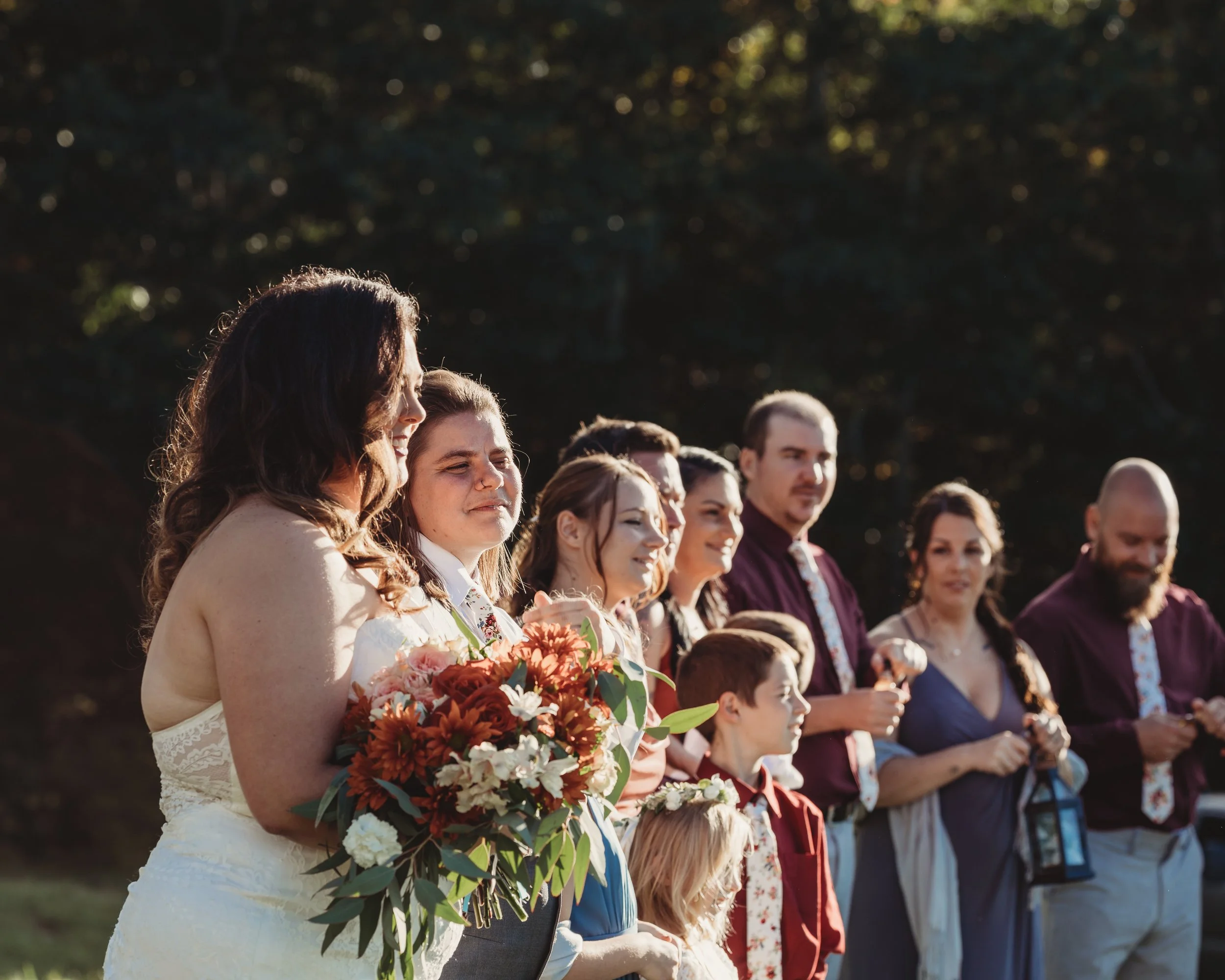 Group of people at an outdoor wedding ceremony, with a bride holding a bouquet of flowers and others dressed in formal attire standing in a line in front of a dark forest background.