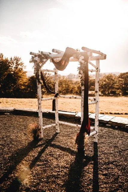 A pair of red and black gymnastic silks hanging from a wooden outdoor gymnastic structure, with a field and trees in the background at sunset.