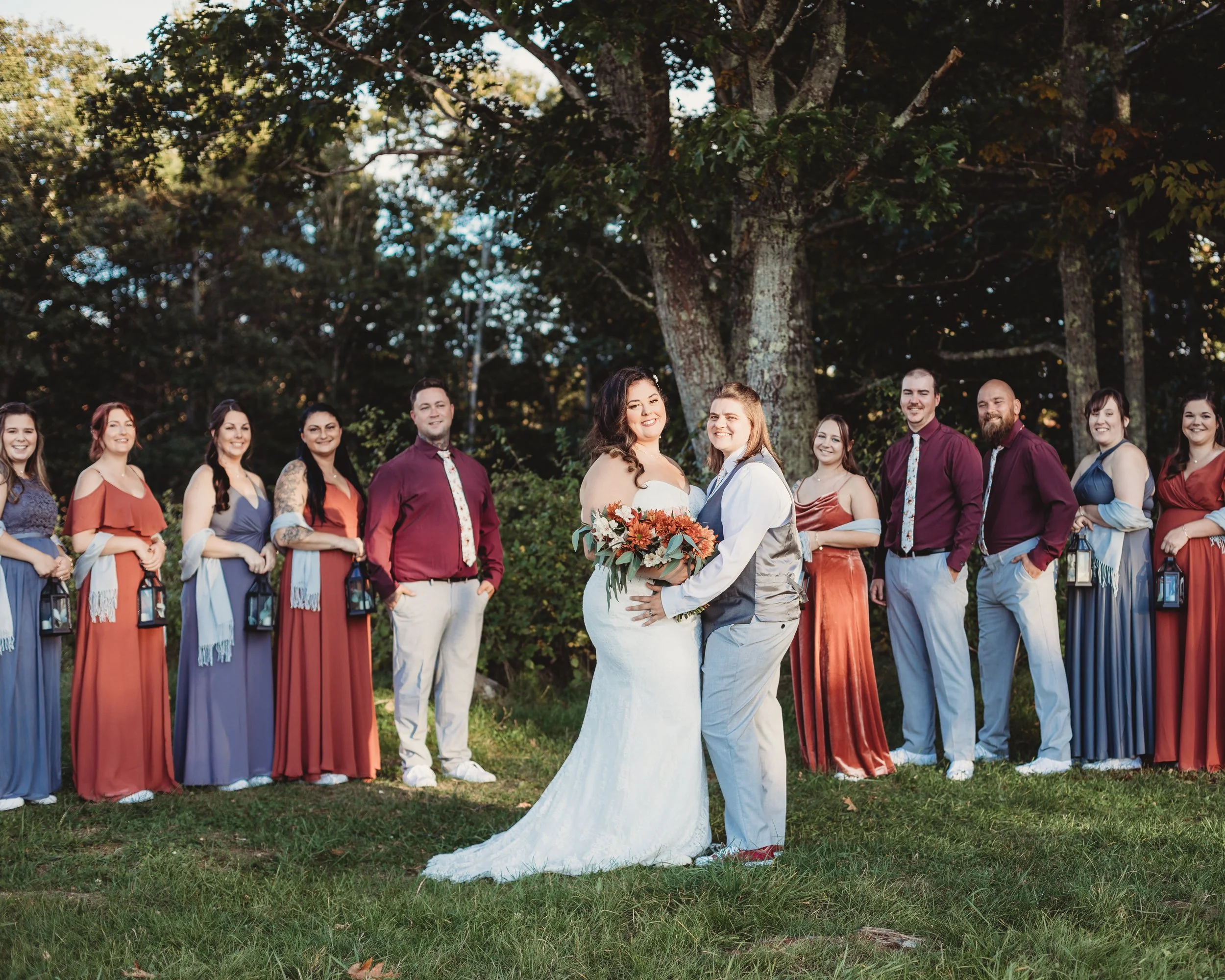 A same-sex wedding ceremony outdoors with a bride in a white gown holding a bouquet and a bride in gray vest and white dress, surrounded by their wedding party in colorful dresses and formal outfits, standing on grass under large trees.