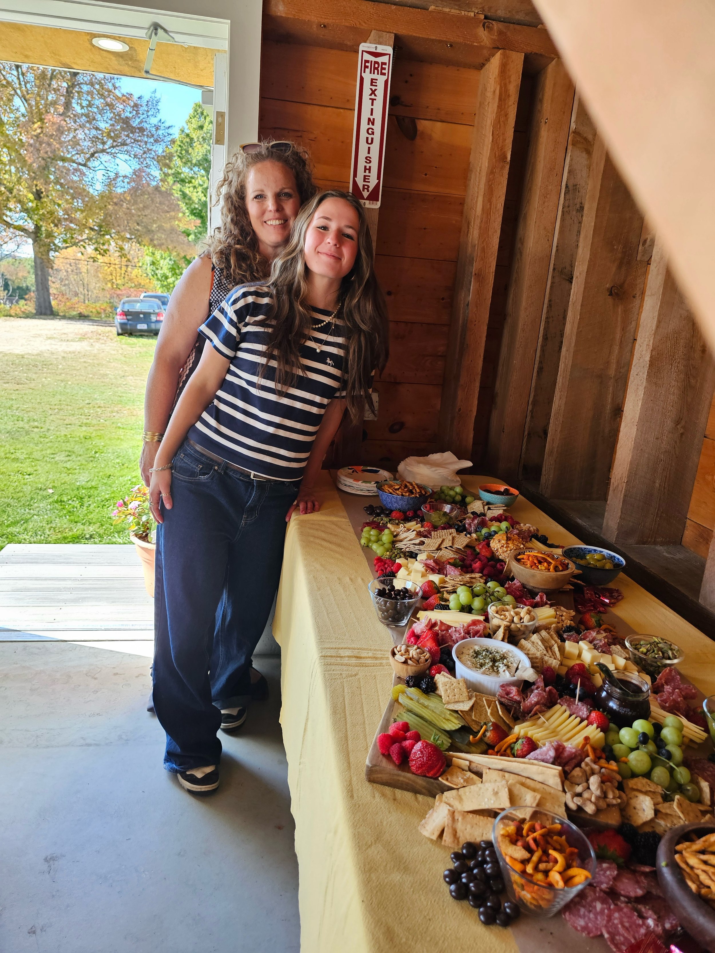 Two women standing beside a table filled with a variety of snacks and appetizers, with a wooden wall and a window showing trees outside.