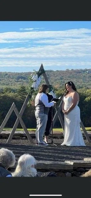 A woman in a white wedding dress standing during an outdoor wedding ceremony, with a officiant reading. The backdrop includes a scenic landscape with trees and a partly cloudy sky.