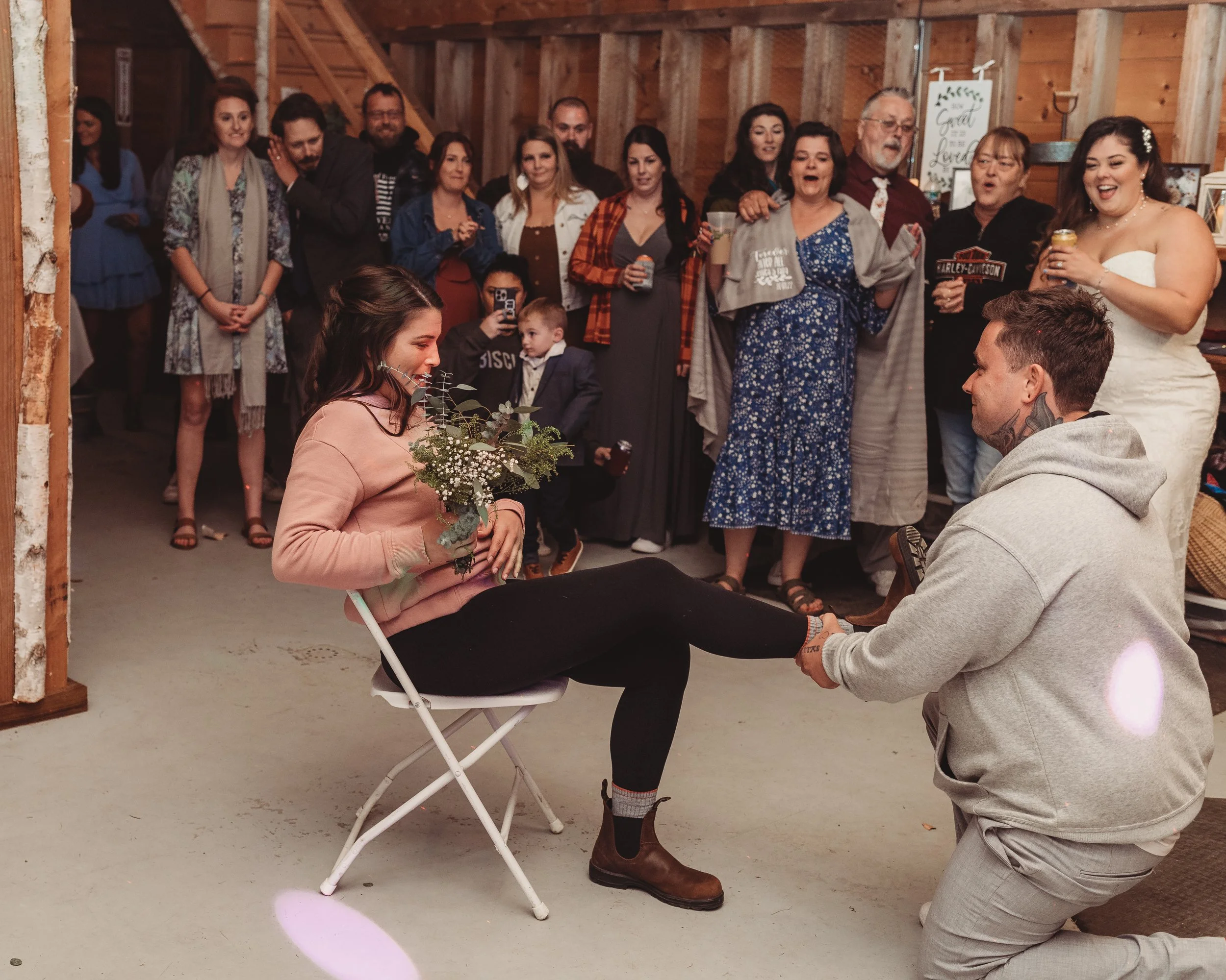 A man on one knee proposes to a woman sitting on a chair holding a bouquet, while a group of friends and family watch and celebrate a wedding or engagement in a rustic indoor setting.
