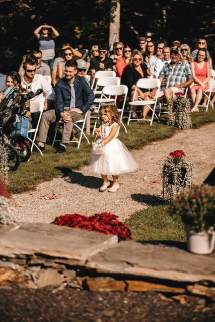 A young girl in a white dress walking down an aisle at an outdoor wedding ceremony, with guests seated on white chairs watching.