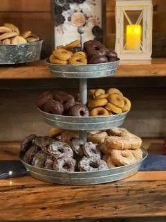 A three-tiered metal serving tray on a wooden surface, filled with assorted donuts including chocolate, glazed, and powdered varieties, with a cozy background featuring a candle and decorative elements.