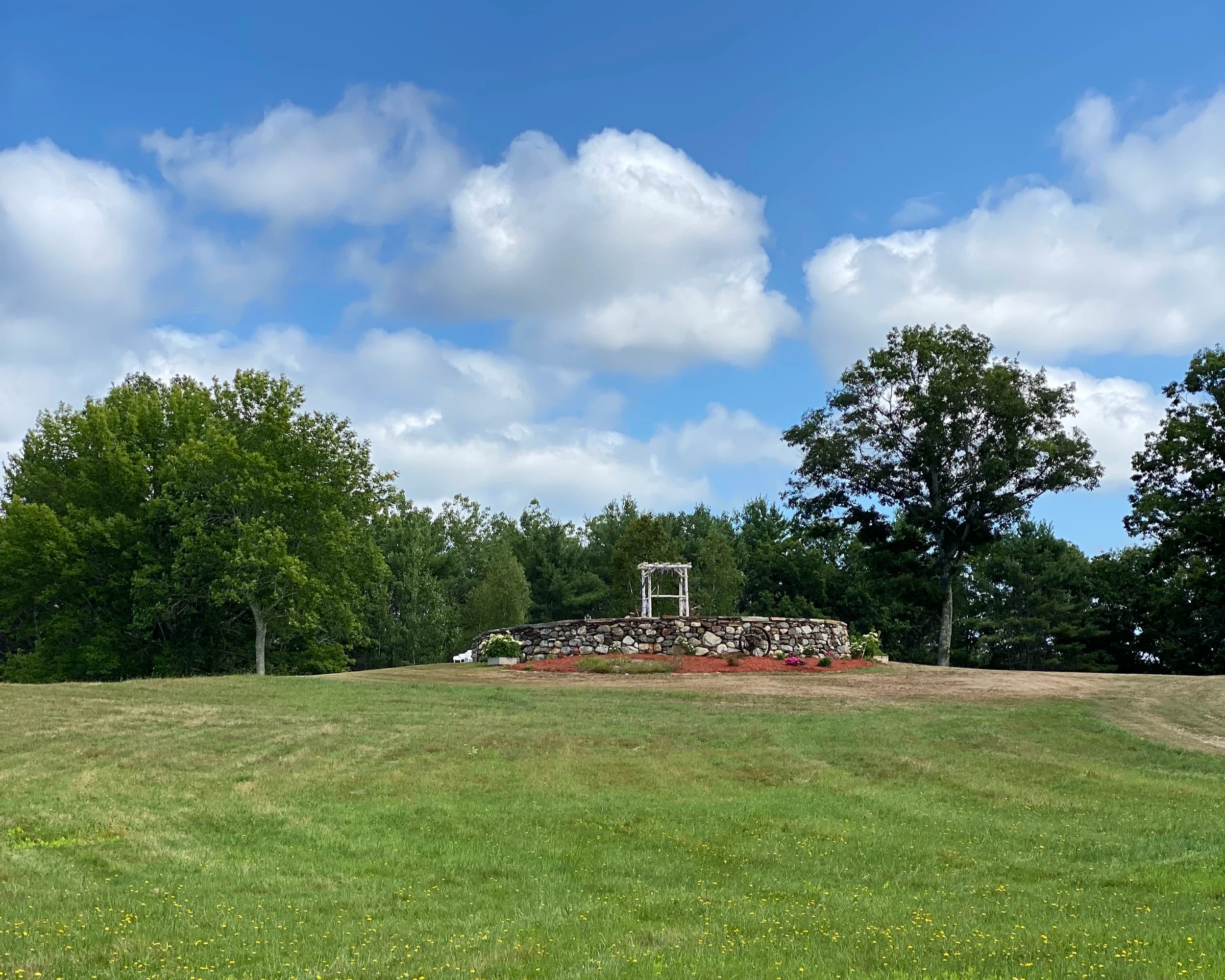 Open grassy area with a stone circle in the distance, trees behind it, and a blue sky with white clouds overhead.