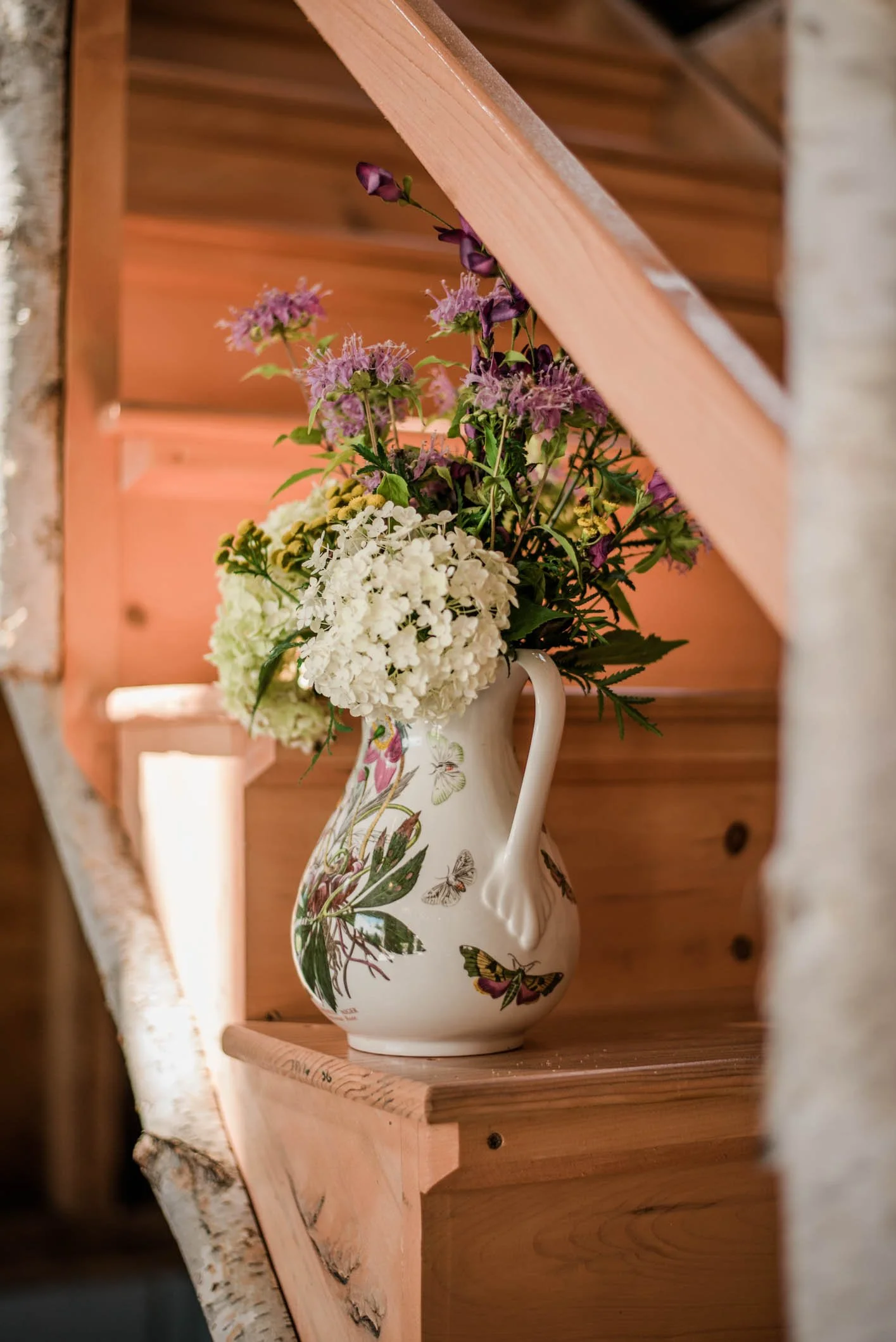 A floral arrangement in a decorative vase with butterflies is placed on a wooden stair step, with a wooden staircase and wall in the background.