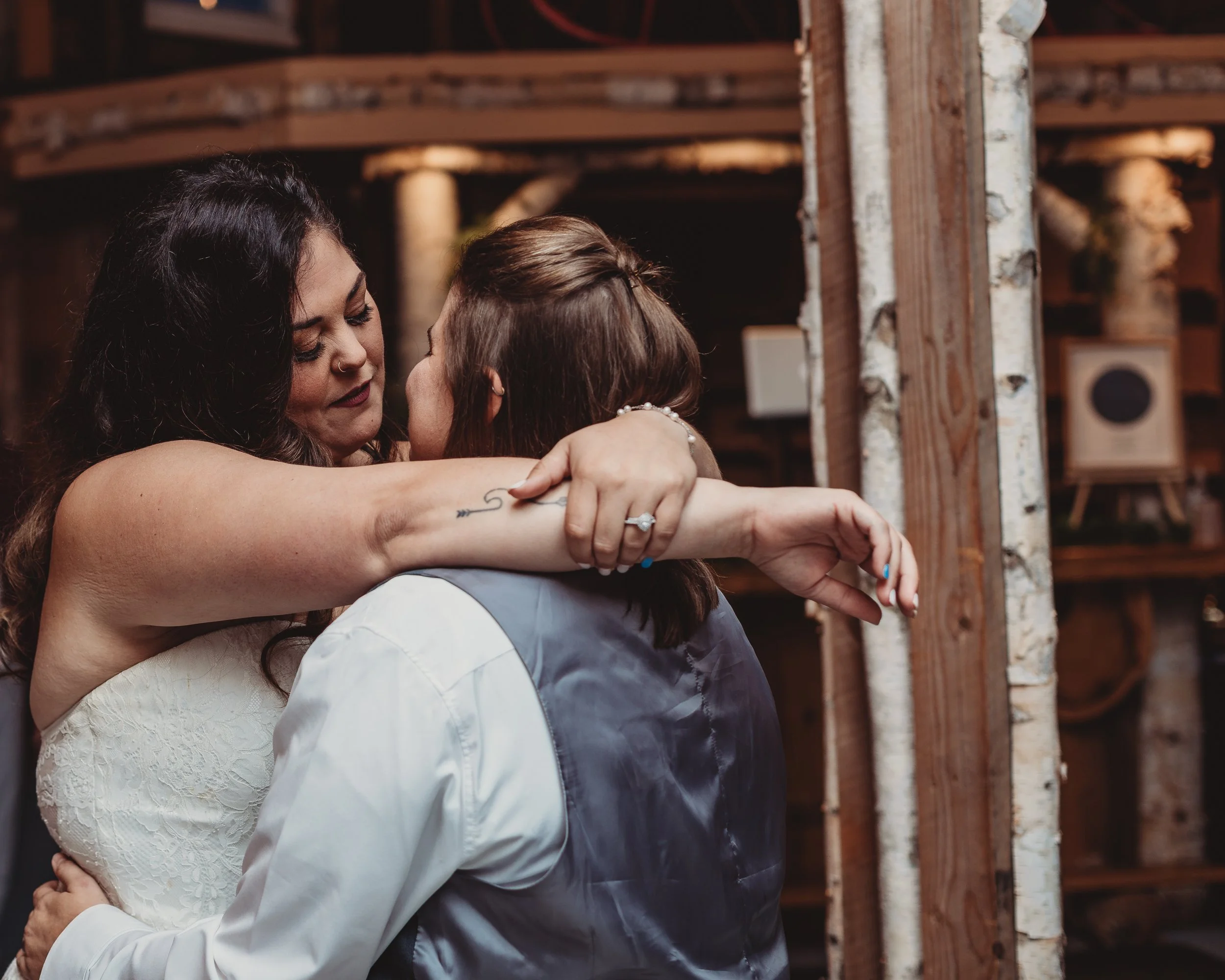 Two women embrace warmly at a rustic indoor event, one in a white lace dress and the other in a gray dress with a satin vest, sharing an intimate moment.