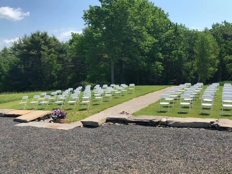 Outdoor wedding ceremony setup with white chairs arranged on grass, a stone pathway in the center, and trees in the background.