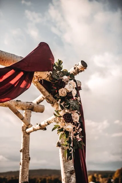 Wedding arch decorated with flowers and burgundy fabric, set outdoors against a cloudy sky.