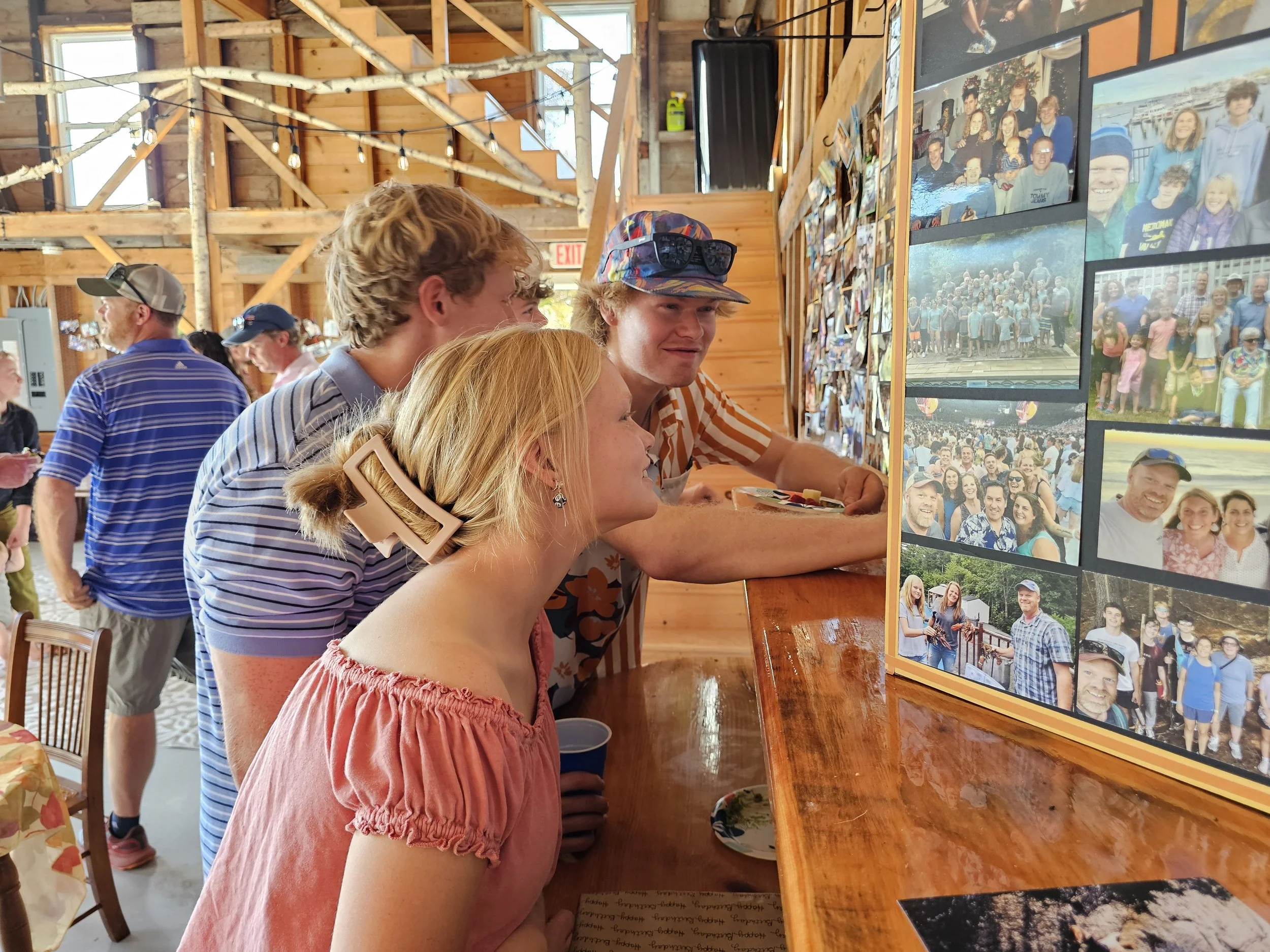 Four people are looking at a collage of group photos on a wooden wall, inside a rustic venue. The group includes a woman with blonde hair and a pink dress, a man with on a floral shirt and sunglasses on his head, and two young men with curly hair, one wearing a striped shirt and the other a cap. The background shows other people, some standing and some at tables, inside the wooden structure.