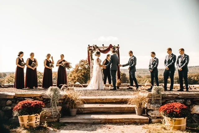 A wedding ceremony taking place outdoors on a stone platform with an arch decorated with flowers. The bride and groom are exchanging vows in the center, surrounded by bridesmaids on the left and groomsmen on the right.