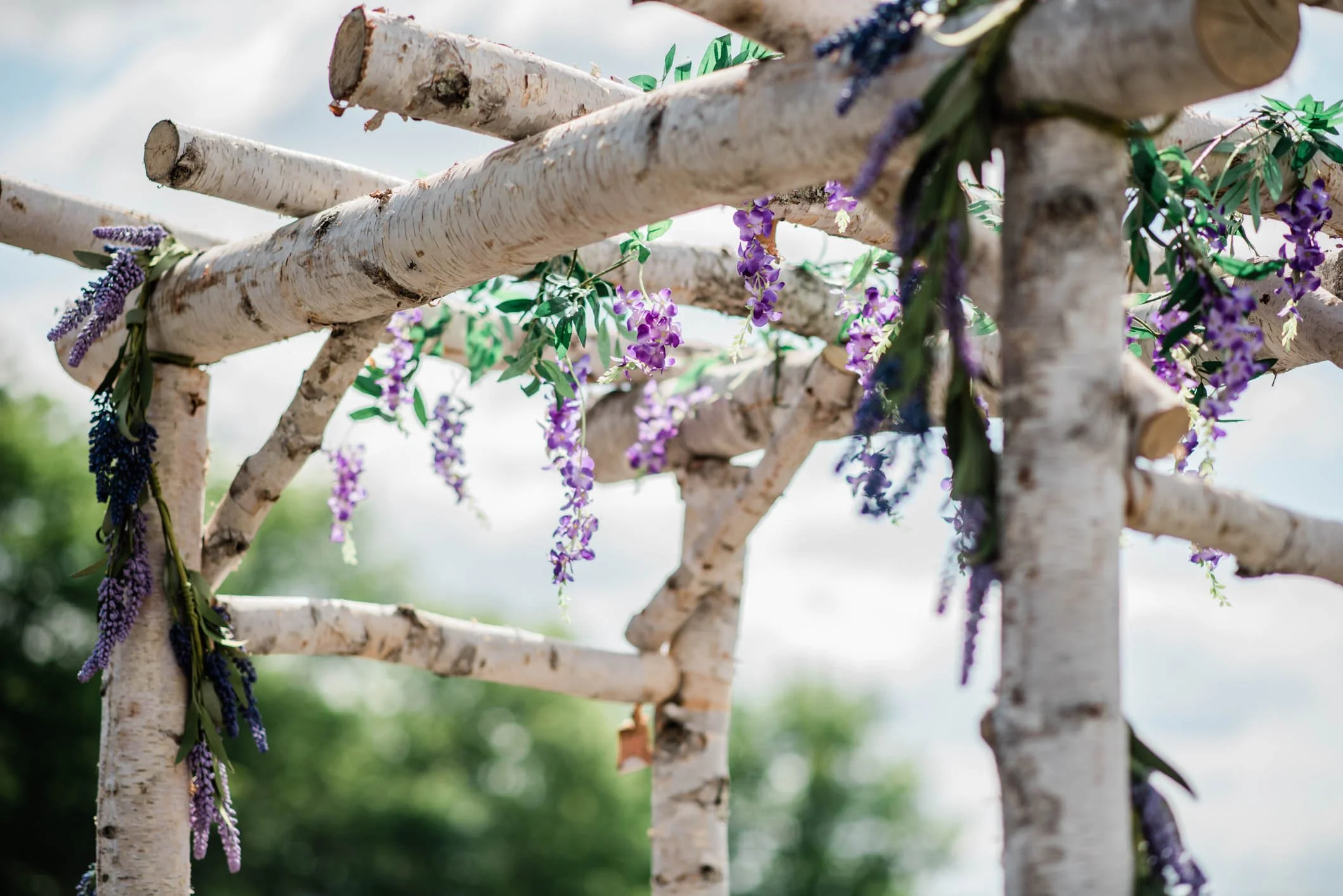 Close-up of a white-painted wooden garden fence decorated with purple and blue artificial flowers and green leaves, with a blurred outdoor background.