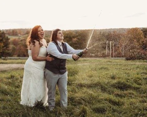 Two women, one in a wedding dress and the other in a suit, laughing and taking a selfie with a selfie stick outdoors in a grassy field during daytime.
