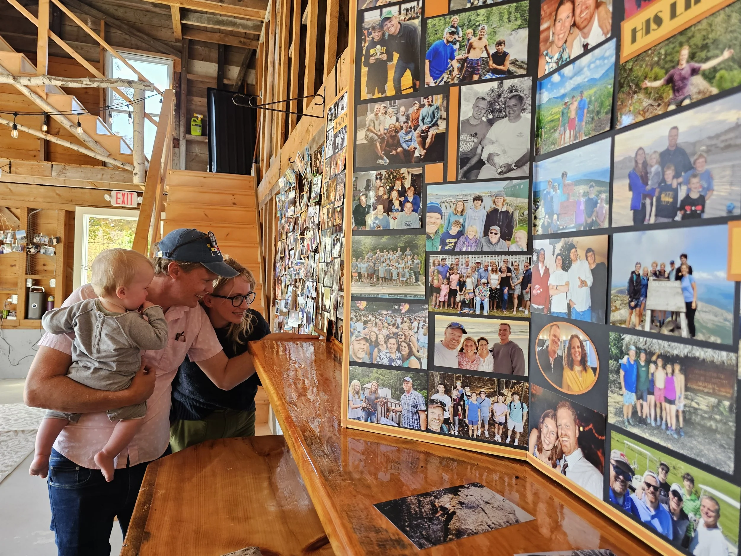 A woman holding a young child, both leaning forward with smiles, looking at a photo display in a rustic wooden room filled with family photos on the wall.
