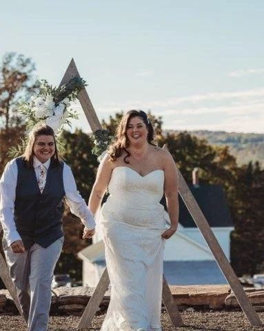 Two women in wedding attire holding hands outdoors with a wooden triangular wedding arch decorated with flowers and greenery in the background.