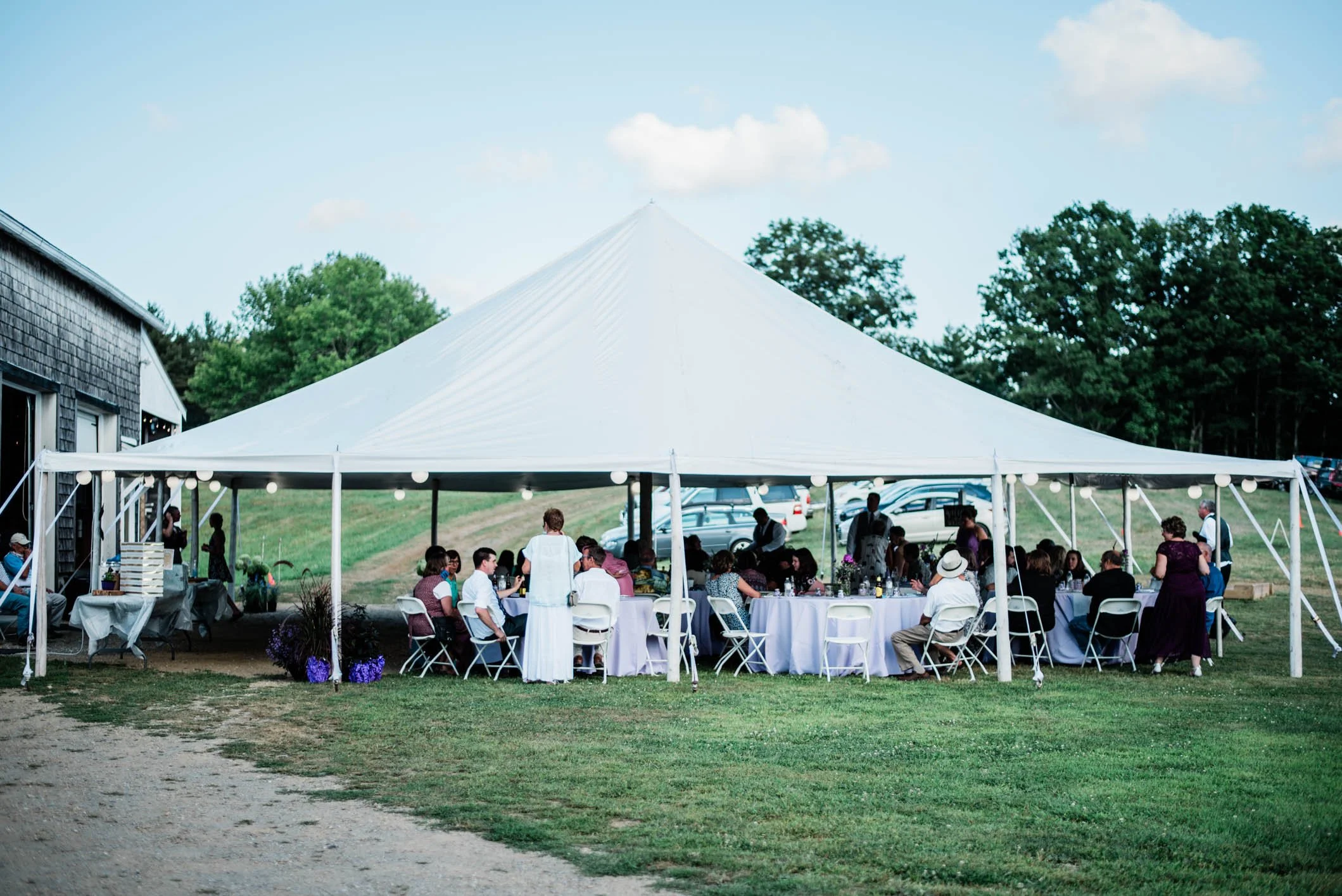 A large outdoor white tent set up for a gathering with people sitting at round tables, some standing and walking, on a grassy area with trees in the background and a partly cloudy sky.