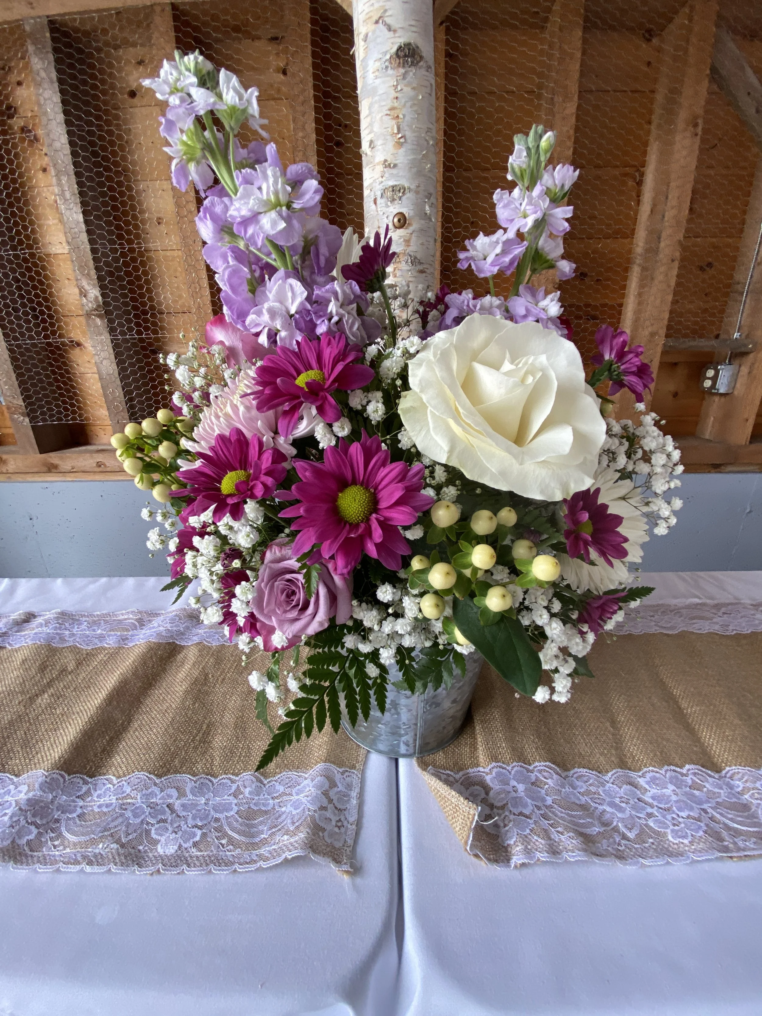 A colorful floral arrangement in a silver metal bucket, including purple, white, and pink flowers, placed on a table with a white tablecloth and burlap runner with lace. The background features a wooden wall and mesh wire.