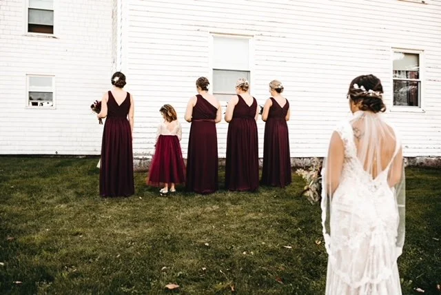 Bride in a white wedding dress and veil looks at five bridesmaids dressed in burgundy gowns standing against a white house exterior.