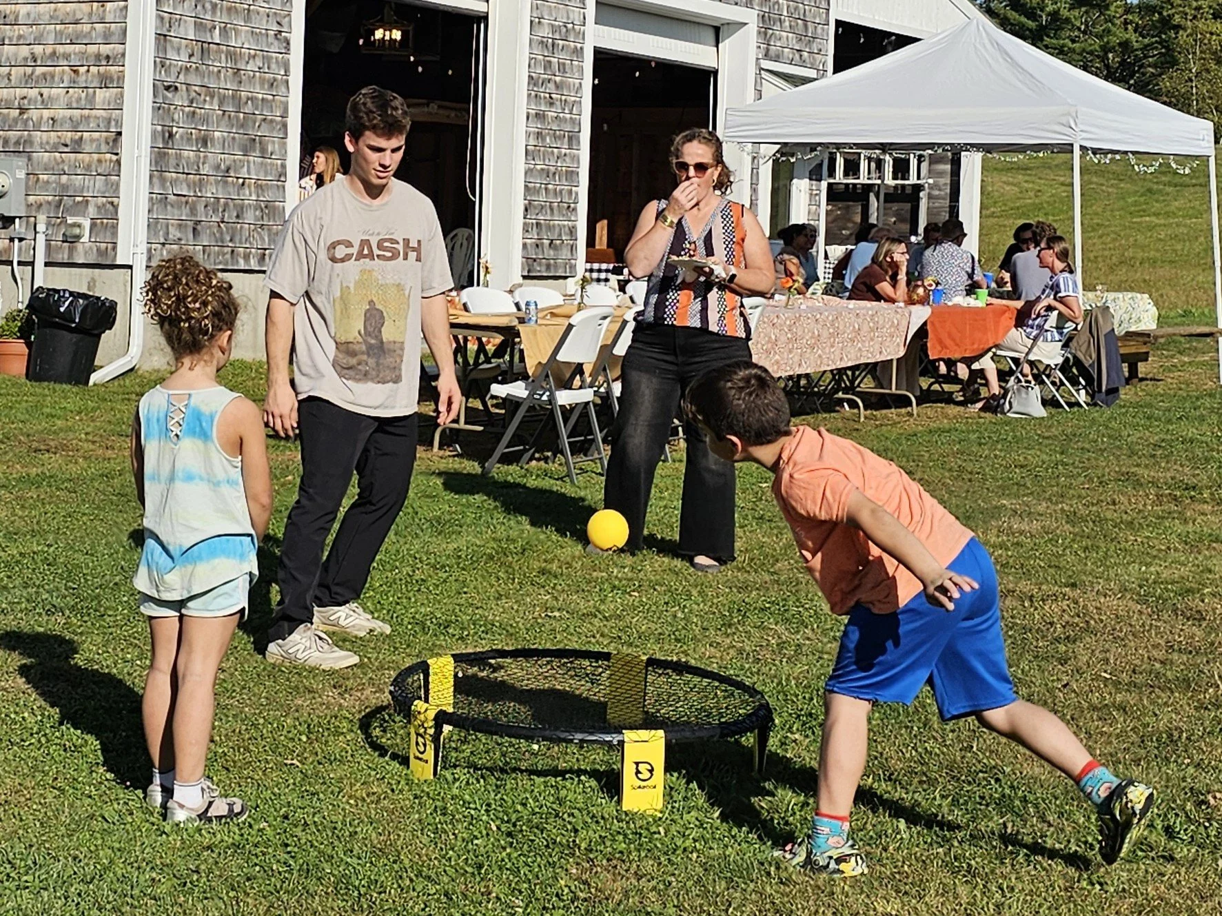 People playing a game of cornhole outside on a lawn during a gathering, with a table of seated guests and a canopy in the background.