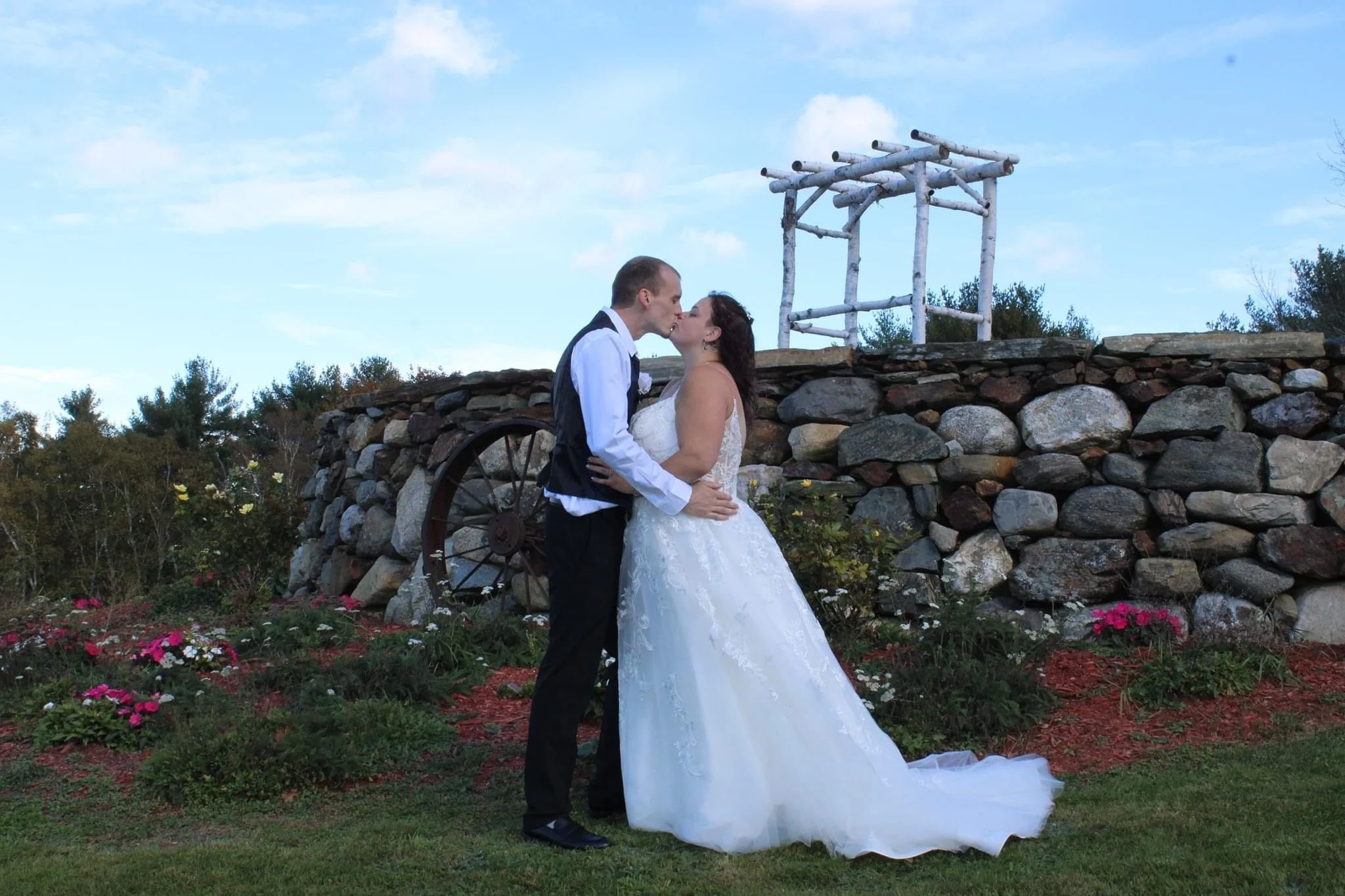 A bride and groom kiss outdoors on a stone wall background with a blue sky, trees, and colorful flowers surrounding them.