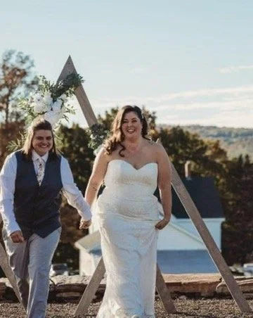 Two women in wedding attire holding hands outdoors, with a wooden wedding arch and a scenic background.