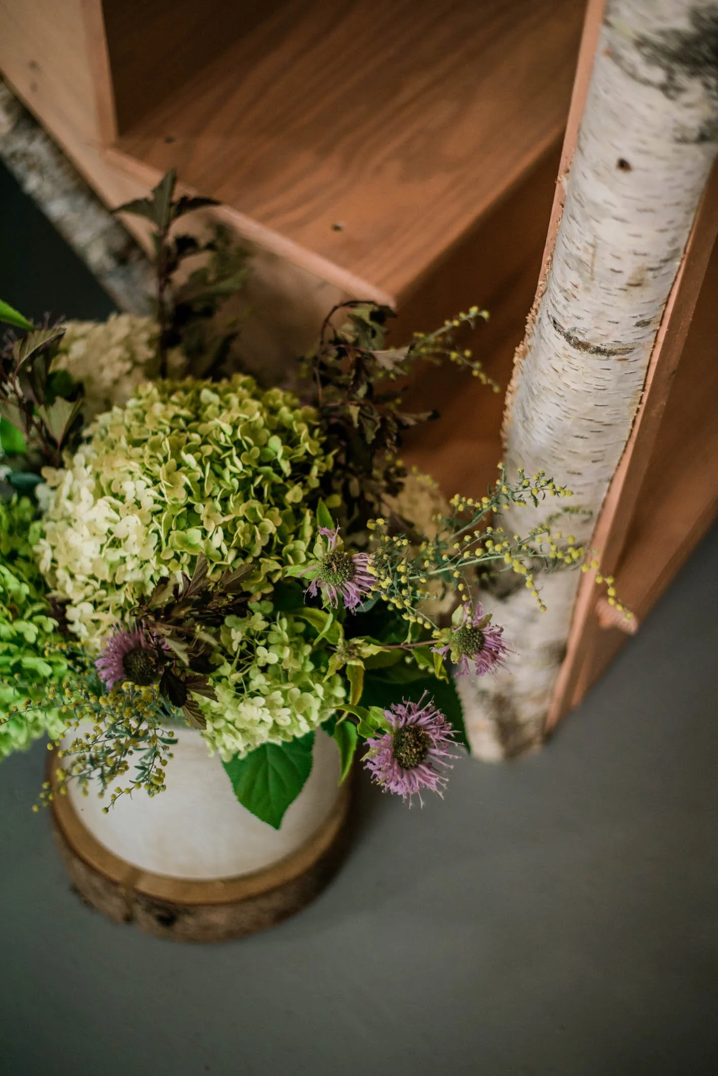 Close-up of a decorative floral arrangement with green hydrangeas, purple flowers, and foliage in a white vase on a wooden slab, next to a birch log and a wooden shelf.