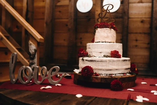 A three-tiered wedding cake with white frosting and red flowers on a wooden table, with a decorative "love" sign beside it, set in a rustic wooden venue.