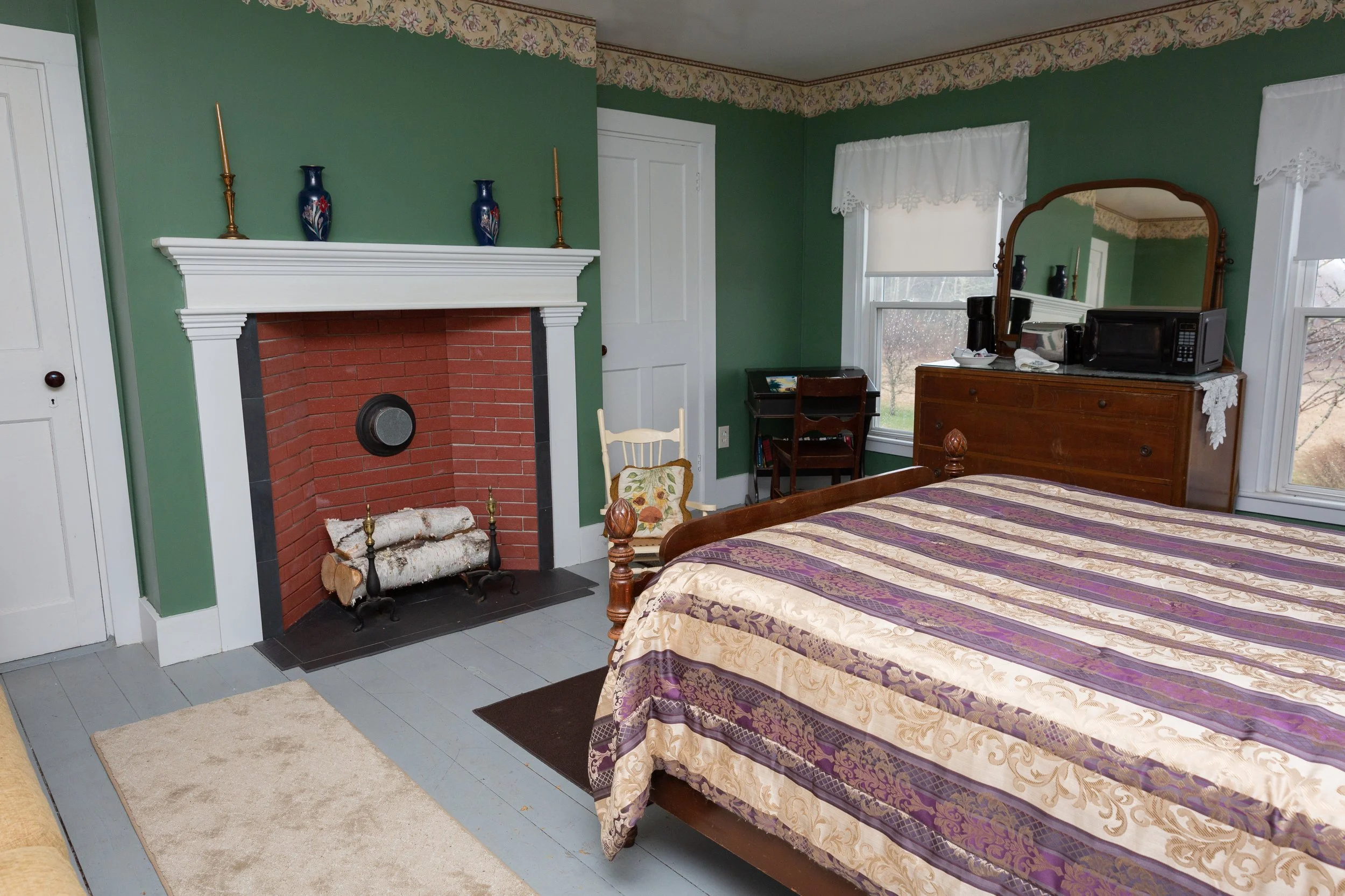 A cozy bedroom with green walls, a fireplace with a white mantel and red brick interior, a bed with a purple and gold patterned quilt, a wooden dresser with a mirror, a small desk with a chair, a white chair with a floral pillow, and a window with wh