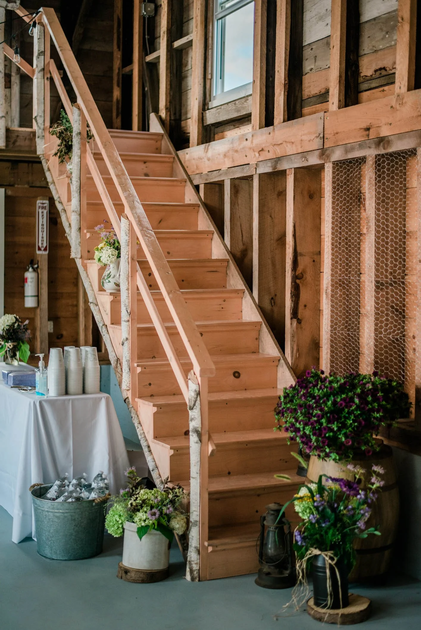 Wooden staircase inside a rustic barn with floral arrangements and a table with cups and drinks nearby.