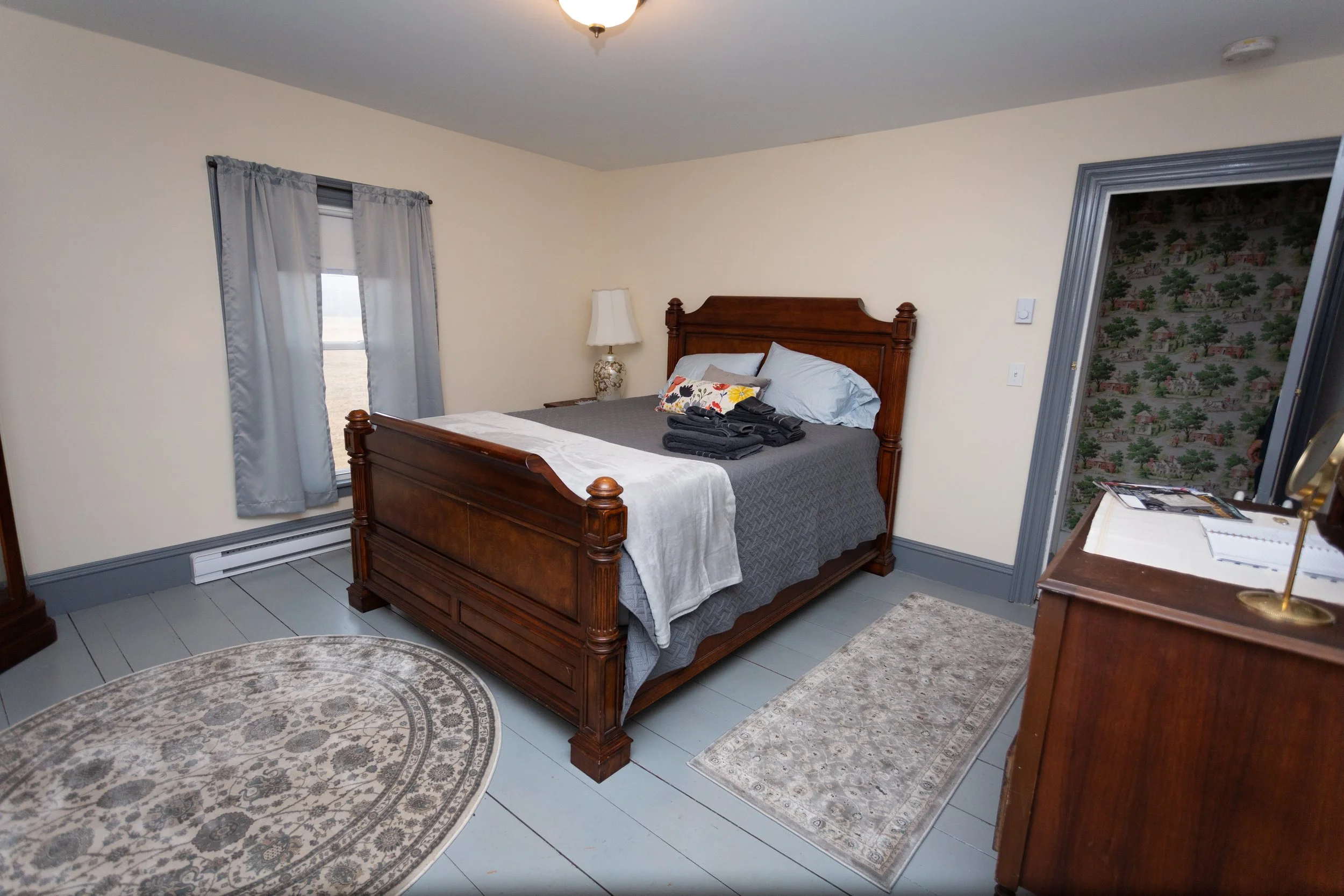 Hotel bedroom with a wooden bed frame, gray bedding, white pillows, a beige lamp, a window with light gray curtains, and a dresser with magazines on top.