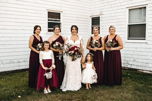 Group of women and children in wedding attire standing on grass outside white building.