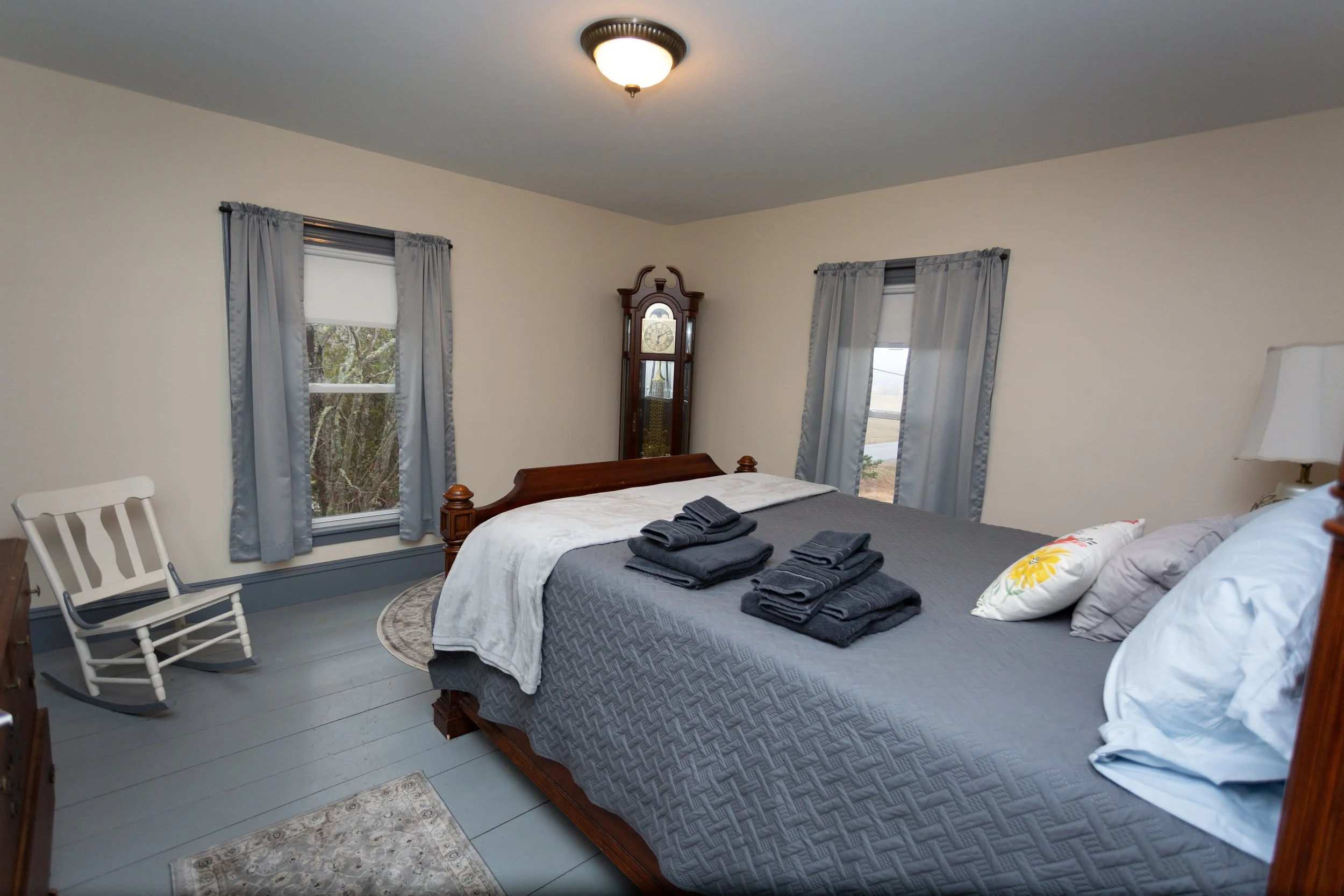 Bedroom with a wooden bed, gray bedding, folded dark towels, and pillows. Two windows with light blue curtains, a white rocking chair, a grandfather clock, a table lamp, and a small rug on a gray wooden floor.