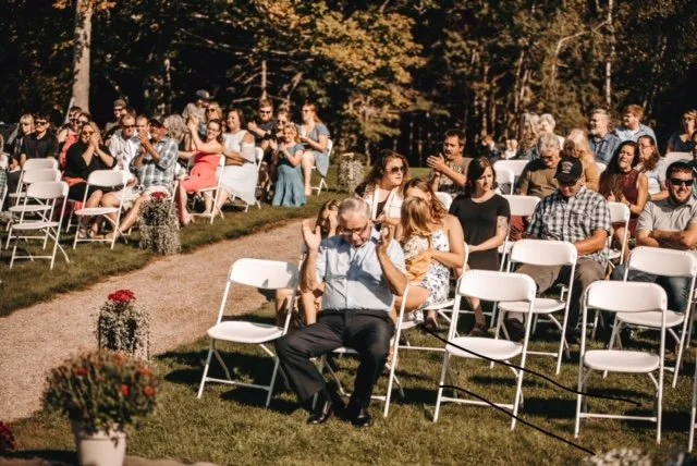 An outdoor gathering with people seated on chairs arranged on grass, some talking and others clapping, during daytime with trees in the background.