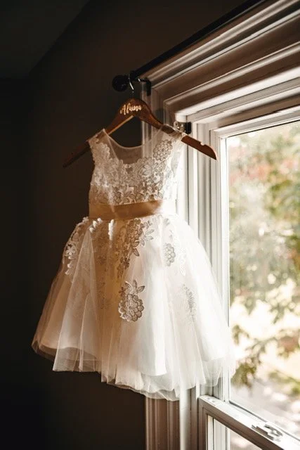 A white lace and tulle girl's dress hanging on a wooden hanger with a name embroidered on it, placed on a ceiling hook near a window with natural light.