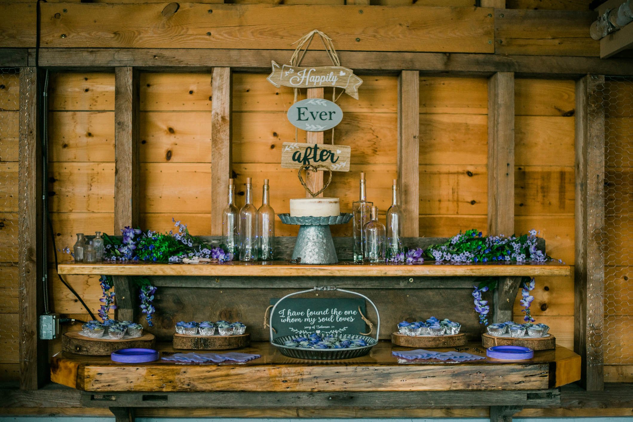 Wedding or celebration dessert table with purple flowers, a cake on a stand, and glasses, against a wooden wall. Hanging sign reads, 'Happily Ever after.'
