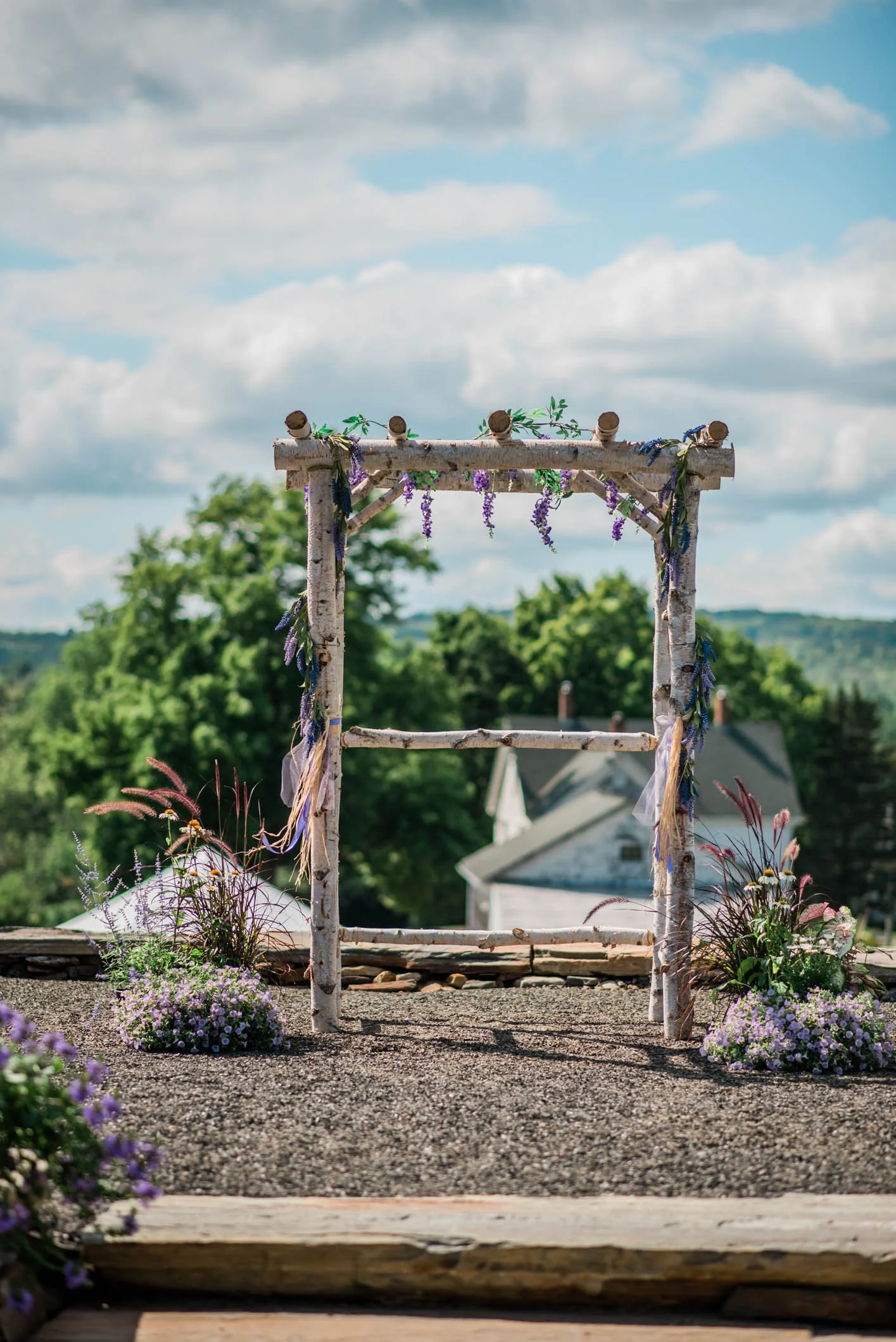 A rustic wedding arch decorated with purple flowers and greenery, standing on a rooftop with a view of trees and houses under a blue sky with clouds.