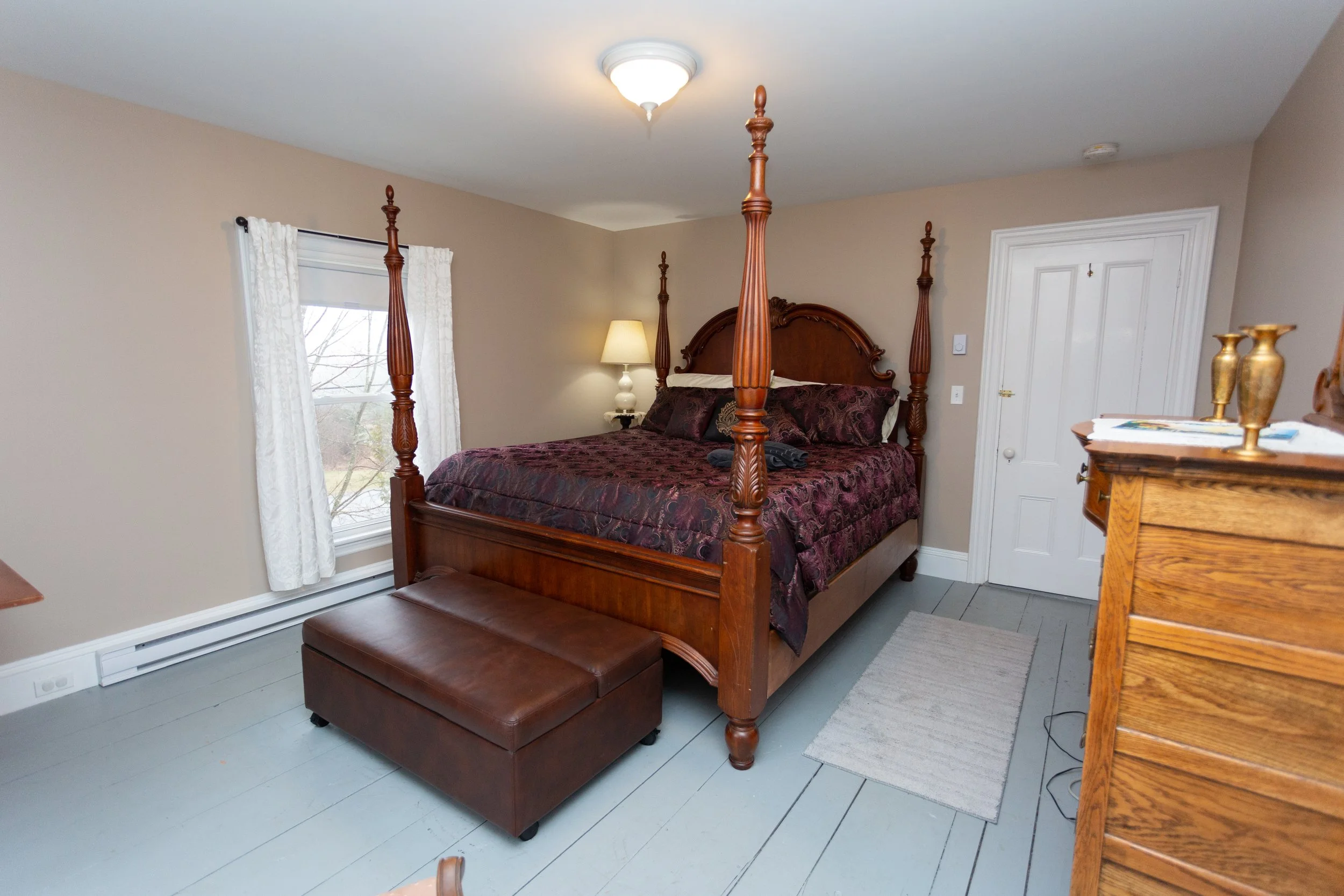 Bedroom with a wooden four-poster bed, dark patterned bedding, a small bedside lamp, a brown leather bench at the foot of the bed, a window with white curtains, a dresser with decorative vases, and a small rug on gray wooden flooring.