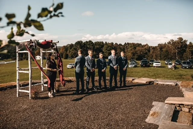 Group of people at an outdoor wedding ceremony with a wooden arch and black gravel ground, parked cars in the background, and trees under a partly cloudy sky.