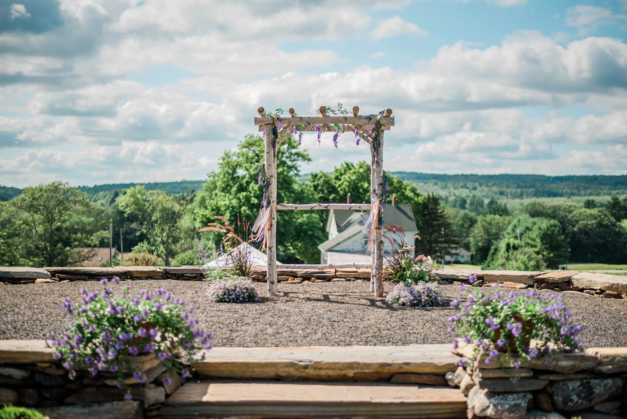 A rustic wedding arch decorated with purple and white flowers, set outdoors on a sunny day with a blue sky and scattered clouds, overlooking a green landscape.
