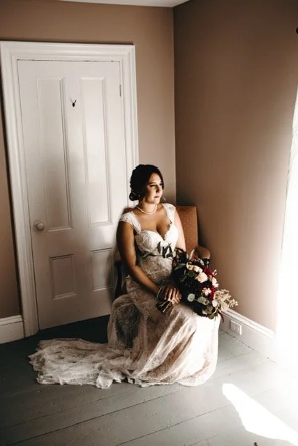 A bride sitting on a chair in a room, holding a bouquet of flowers, wearing a white wedding dress with lace details, with a closed door behind her and sunlight coming through a window.