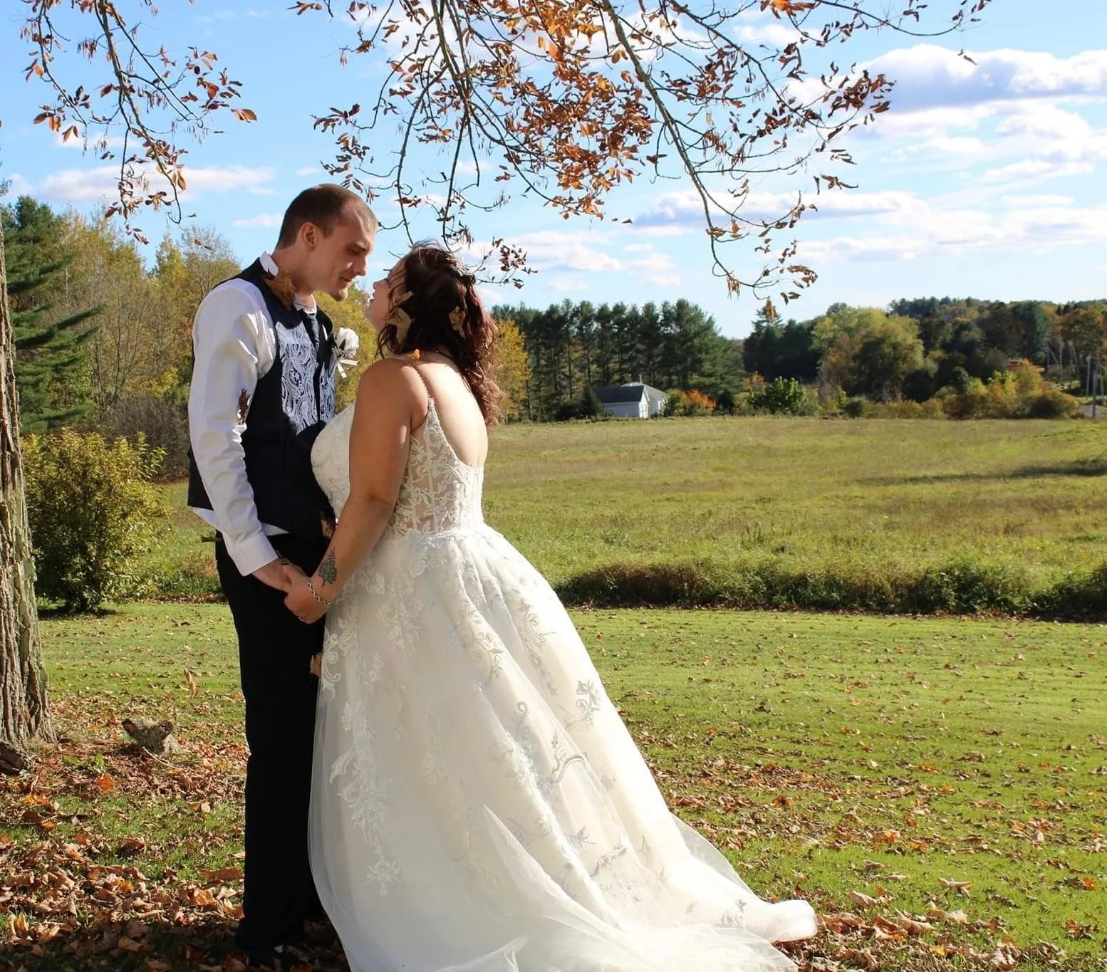 A bride and groom share a loving moment outdoors on a fall day in a grassy field with trees and a small building in the background, under a partly cloudy sky.