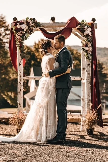 Bride and groom kissing under a floral and fabric decorated arch at their wedding outdoors during sunset.