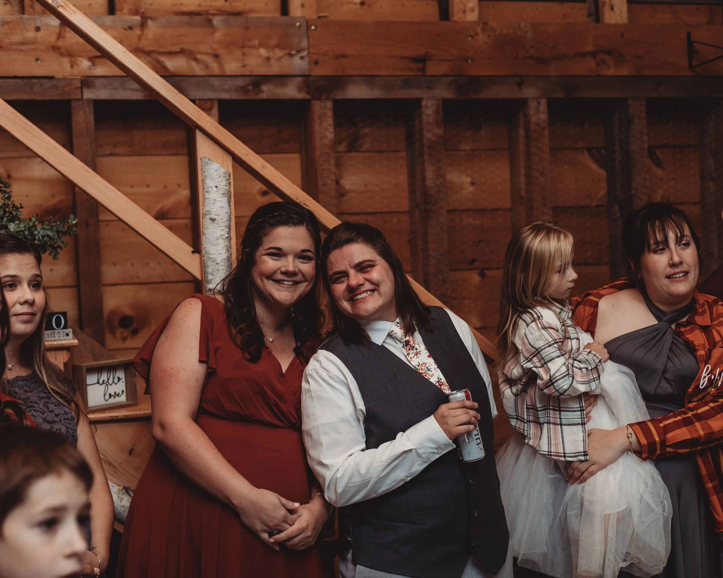 Group of people, including a smiling woman in a red dress, a man in a vest holding a beverage, and a woman holding a young girl, gathered indoors with wooden walls and staircase in the background.