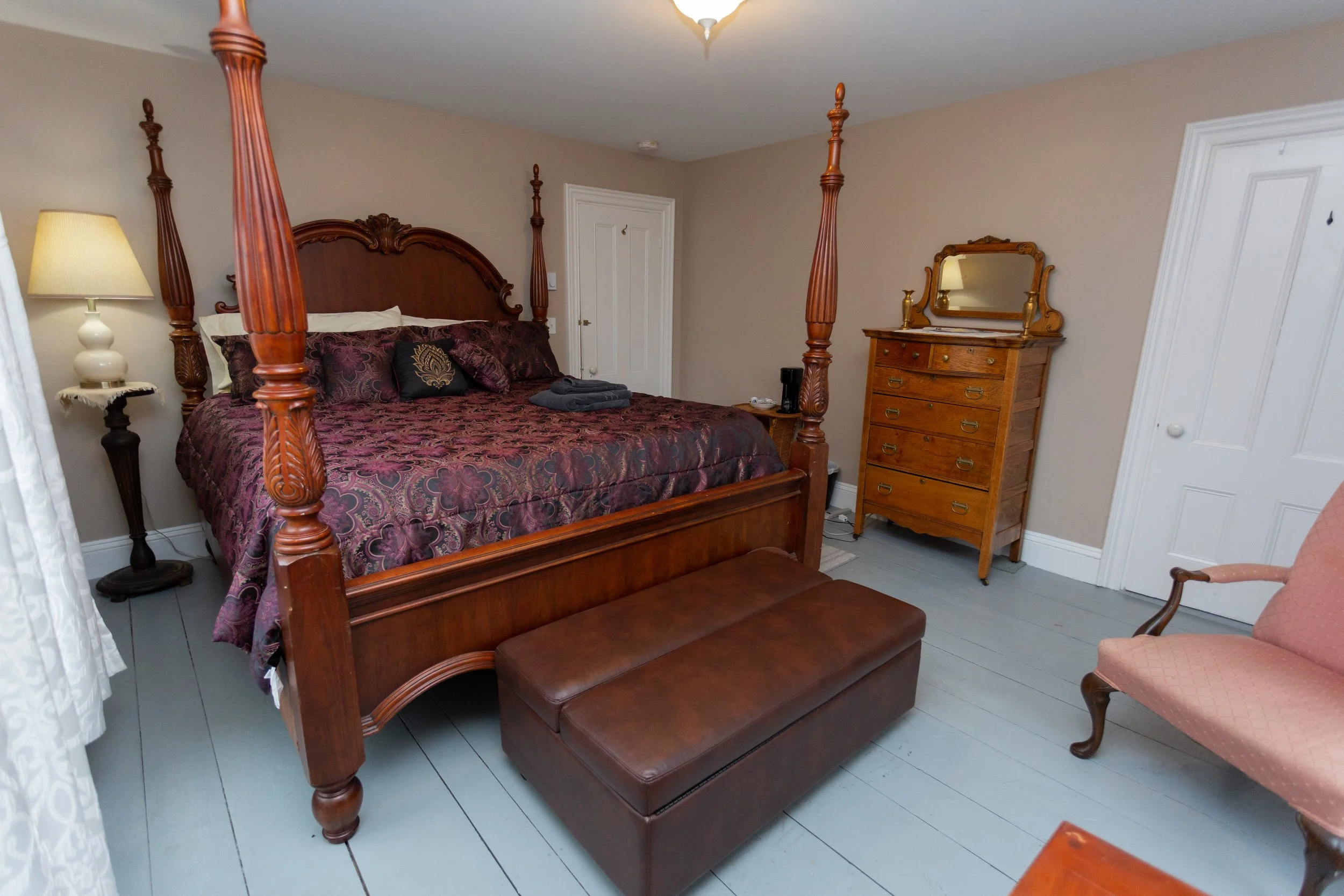 A bedroom with a four-poster bed, brown and purple bedding, a leather bench at the foot of the bed, a pink upholstered armchair, a wooden dresser with a mirror, a side table with a lamp, and some folded towels on the bed.