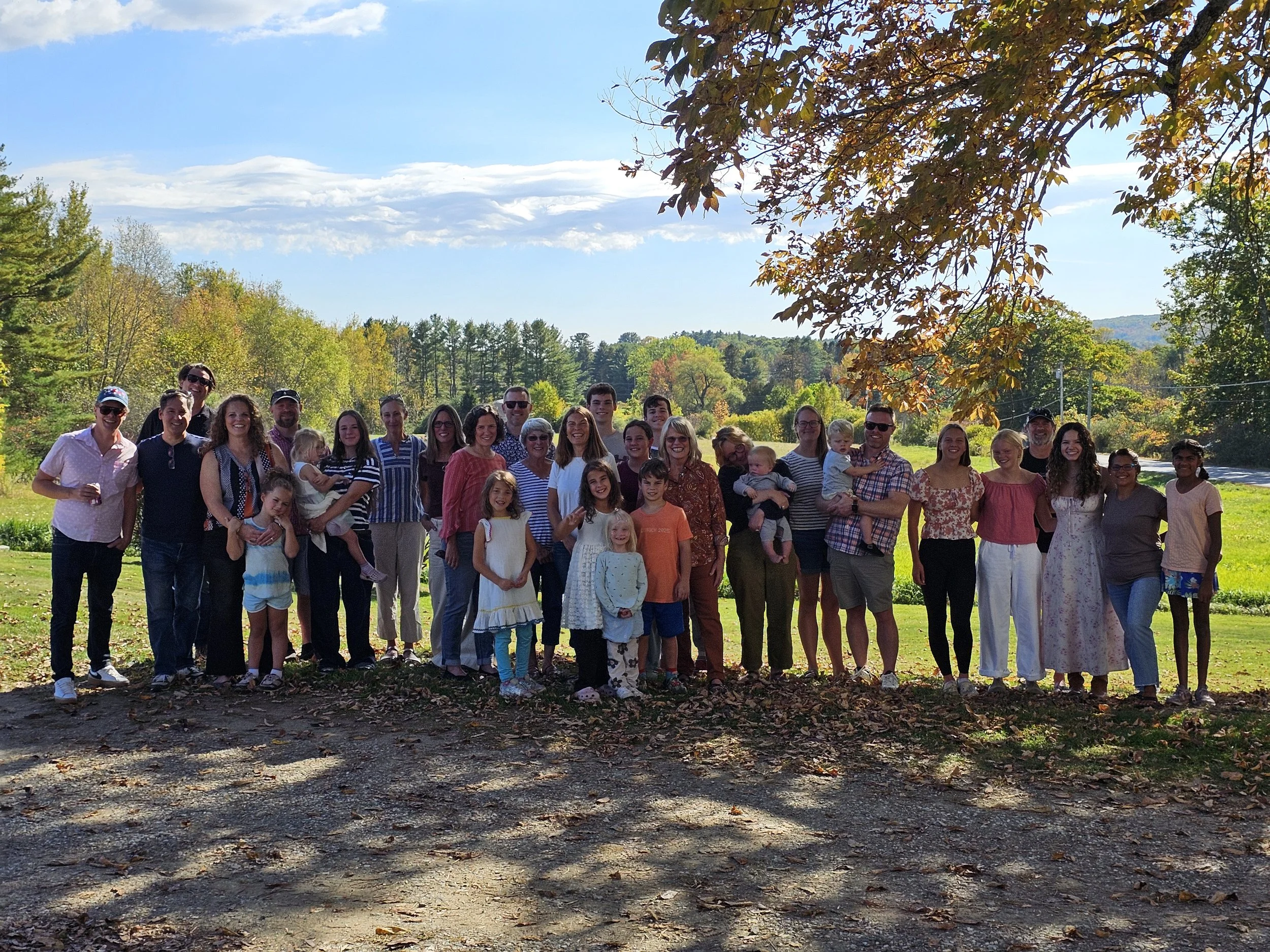 A large group of people, including children and adults, standing outdoors under a tree with autumn leaves, on a sunny day with a background of green trees and blue sky.
