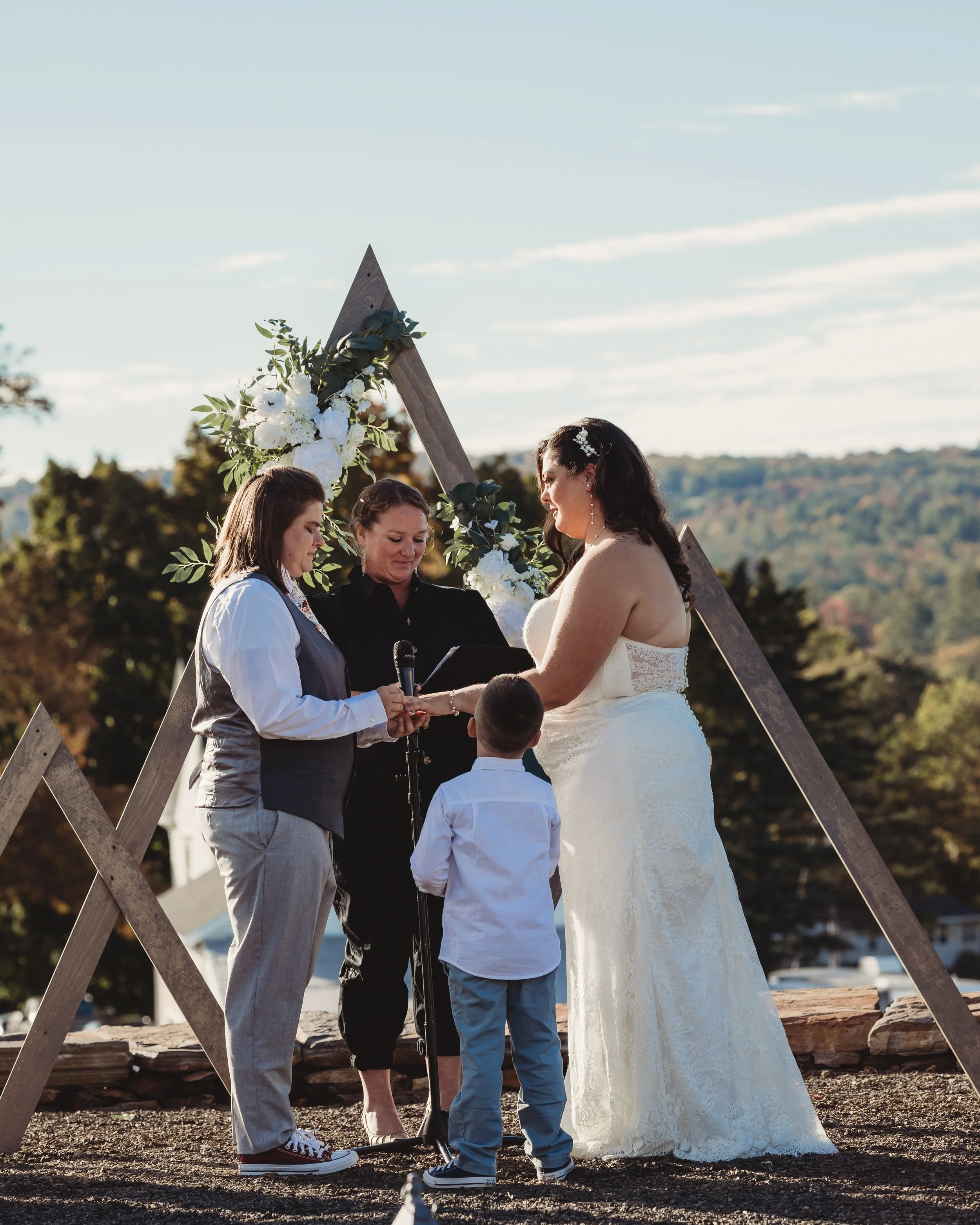 A wedding ceremony taking place outdoors with a bride, groom, officiant, and a young boy, under a wooden triangular arch decorated with white flowers and green leaves, in a scenic setting with trees and hills in the background.